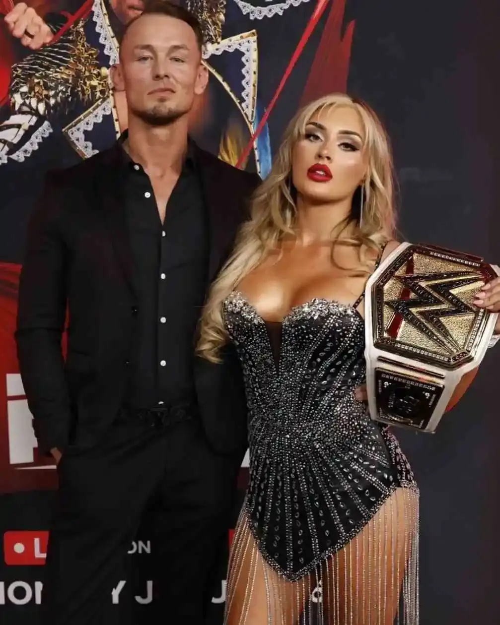 Tiffany Stratton poses with Ludwig Kaiser in a black suit, where she is holding a championship belt and wearing a sparkling black dress with fringe.
