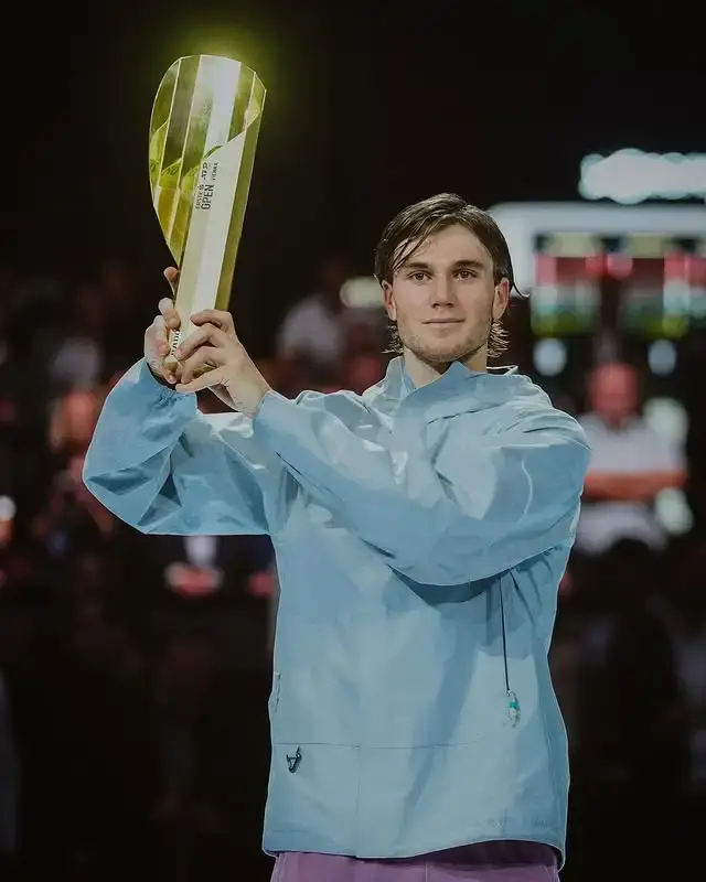 Jack Draper, a male tennis player, holds a gold, curved ATP 500 trophy overhead on a dark court.