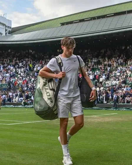Jack Draper walks off a grass court after a match, carrying two tennis bags over his shoulders, with a crowd and the stadium visible in the background.