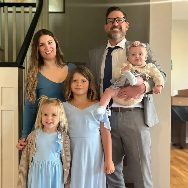 Peter Nottoli smiles in a gray suit and tie, holding a baby with a floral headband, standing with his wife and three daughters, all dressed in light blue, for a family photo.