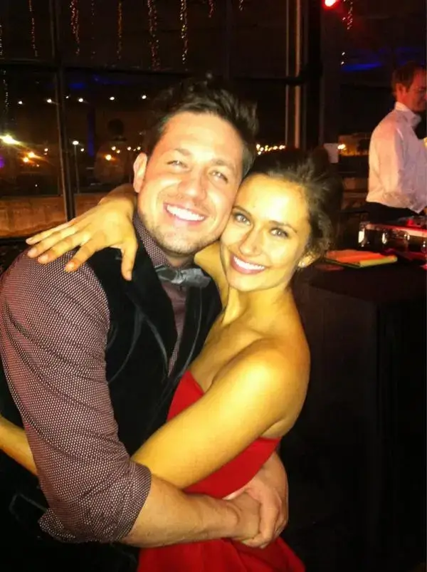 Peter Nottoli and actress Crystal Reed pose closely, smiling at the camera; he is in a vest and patterned shirt, and she is in a red strapless dress.