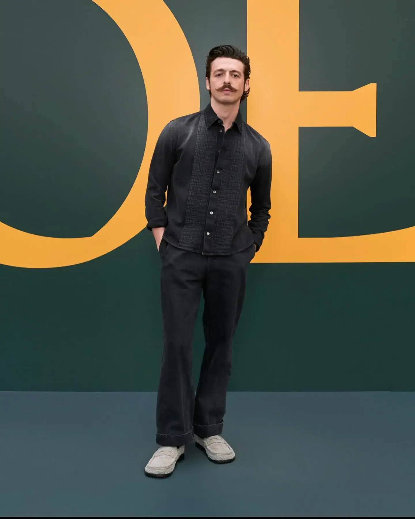 Anthony Boyle, sporting a mustache, wears a textured black shirt and wide-legged trousers with light woven shoes, standing against a dark green and orange graphic background.