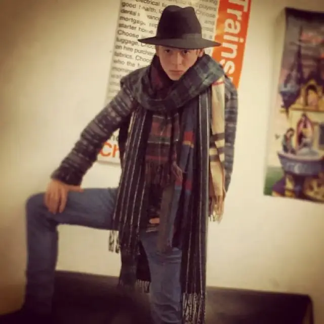 Young Anthony Boyle posing in a dark fedora, a long dark coat, multiple plaid scarves, and jeans, with one leg elevated.