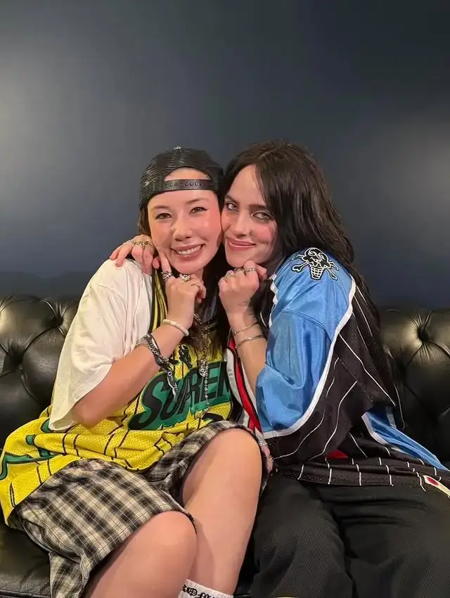 Riisa Naka and Billie Eilish pose for a close photo, both wearing oversized jerseys, with Riisa Naka wearing a backwards cap.