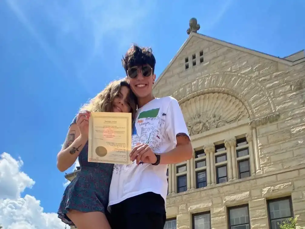 Calysta Bevier and Shawn M. Corchaeo,stand outside a large stone building on a sunny day, holding up a marriage license