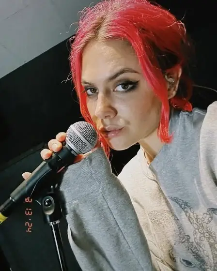 Calysta Bevier in a studio, holding a microphone, wearing a gray sweatshirt and sporting bright pink hair.