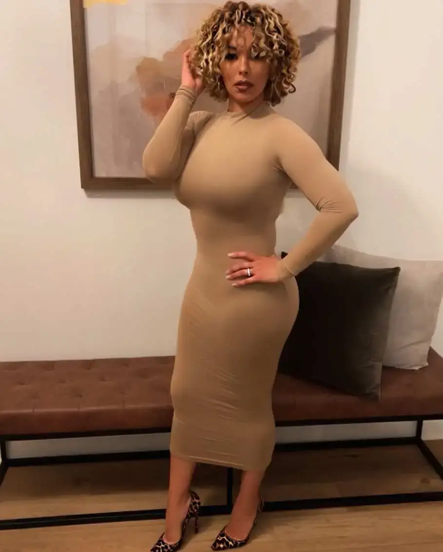 Gloria Govan posing indoors in a figure-hugging beige dress and leopard-print heels.