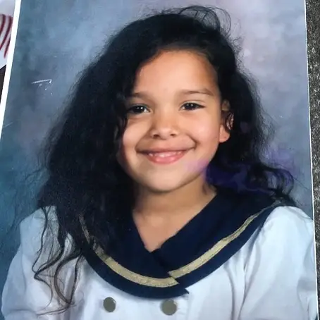 A young Gloria Govan smiling in a school photo, wearing a white outfit with a navy blue sailor-style collar.