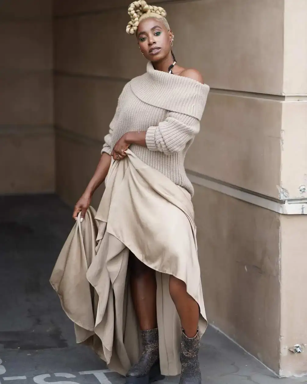 Kirby Howell-Baptiste wearing a beige off-the-shoulder sweater, a flowing tan skirt, and dark boots, with her blonde hair styled in sculptural buns.