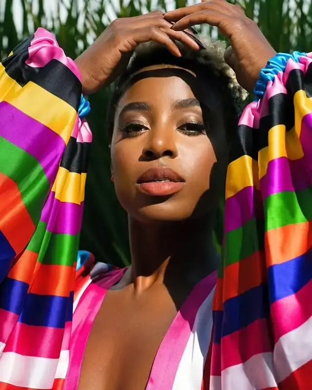 Kirby Howell-Baptiste in a colorful, striped outfit with voluminous sleeves, holding her hands over her head.