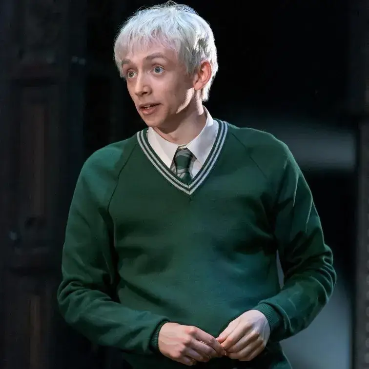 Samuel Blenkin in character with white hair, wearing a green V-neck sweater over a white shirt and striped tie.