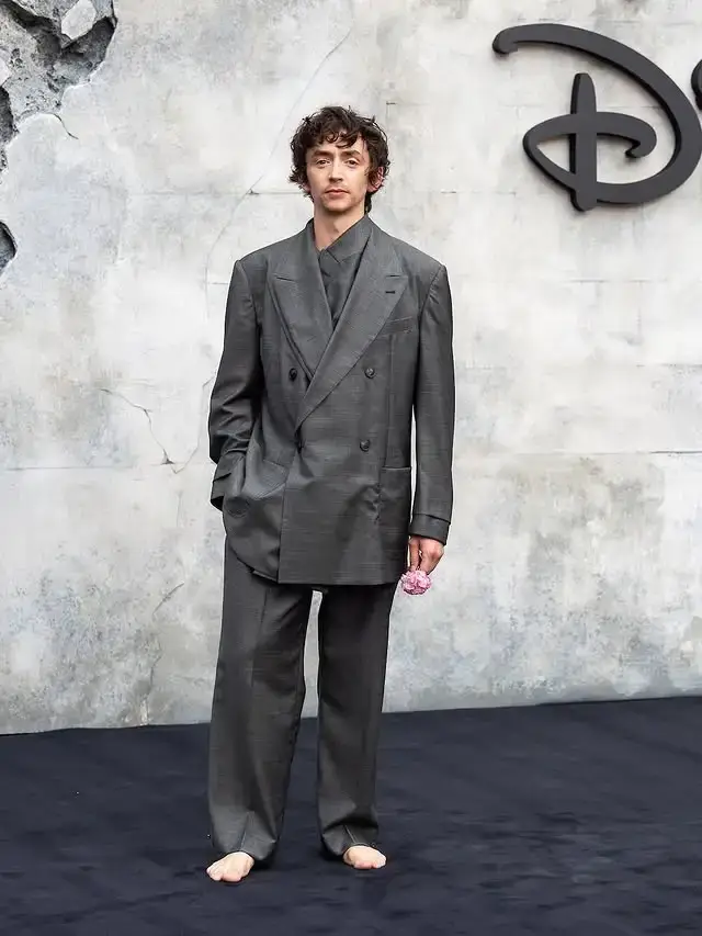 Samuel Blenkin barefoot, wearing an oversized grey double-breasted suit and holding a small pink flower in Disney event.