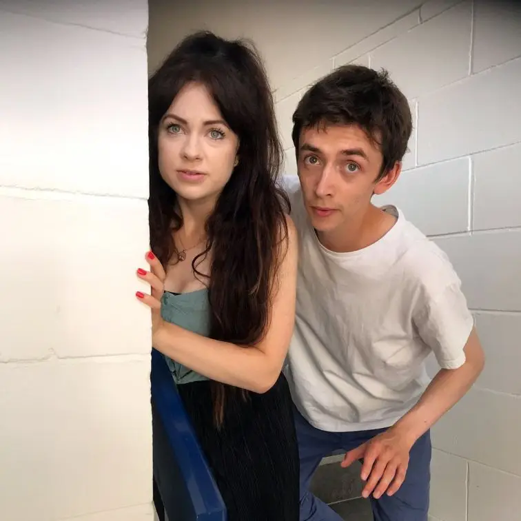 Samuel Blenkin and Lauren Cornelius peeking from around a white wall in a casual.