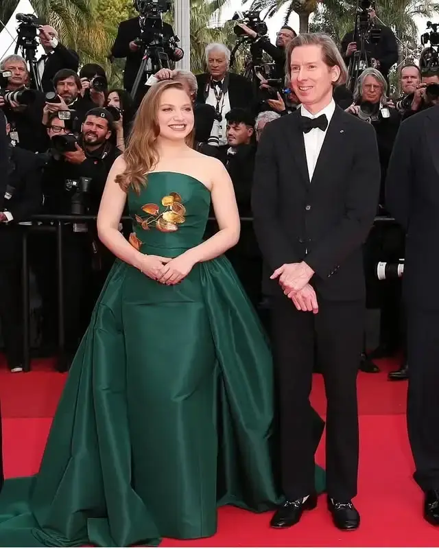 Wes Anderson in a black tuxedo and Mia Threapleton in a long, strapless green gown with a floral design on a red carpet.