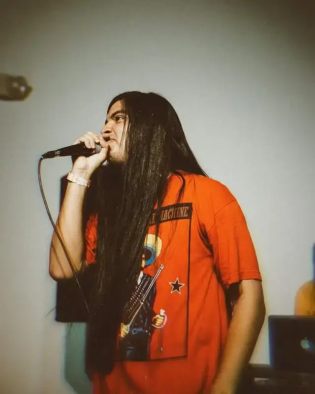 Eduardo Franco with long dark hair, wearing an orange graphic t-shirt, singing passionately into a microphone.