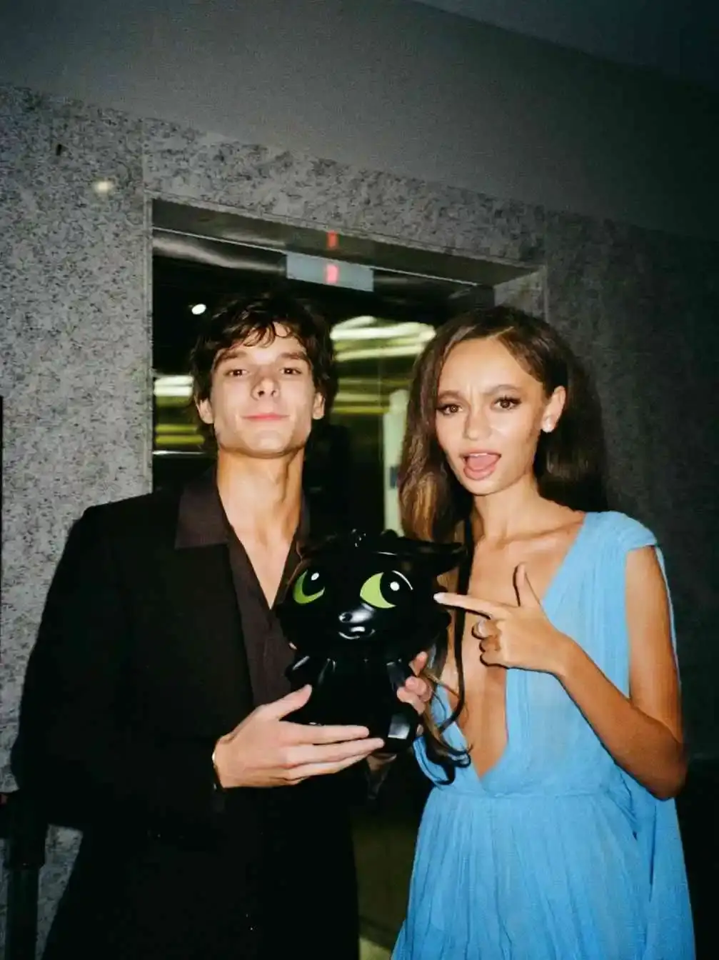 Mason Thames and Nico Parker posing indoors, holding a black dragon-shaped bag, with Parker pointing at it playfully.