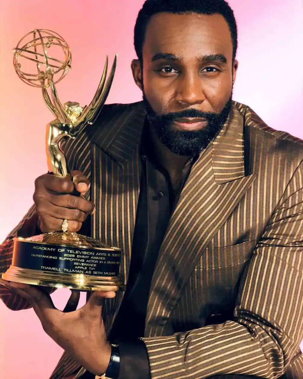 Tramell Tillman, a bearded man in a striped suit, holding an Emmy award and looking at the camera.
