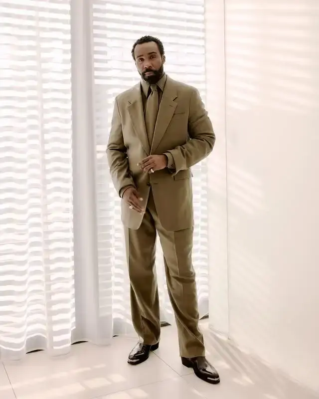 Tramell Tillman in a monochromatic tan suit, standing in front of a window with blinds.