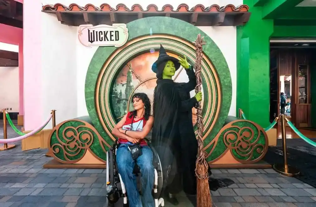 Actress Marissa Bode sits in a wheelchair next to a life-sized statue of the Wicked Witch of the West at a Wicked theme park display.