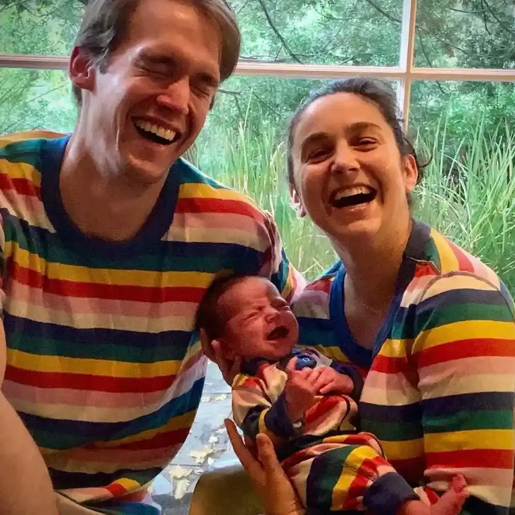 Molly Ephraim and her husband Evan laughing while holding their newborn daughter Zia in matching rainbow-striped outfits.