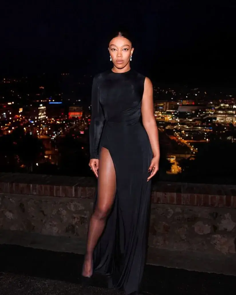 Naomi Ackie posing at night in a long black one-shoulder dress with a high leg slit against a backdrop of city lights.