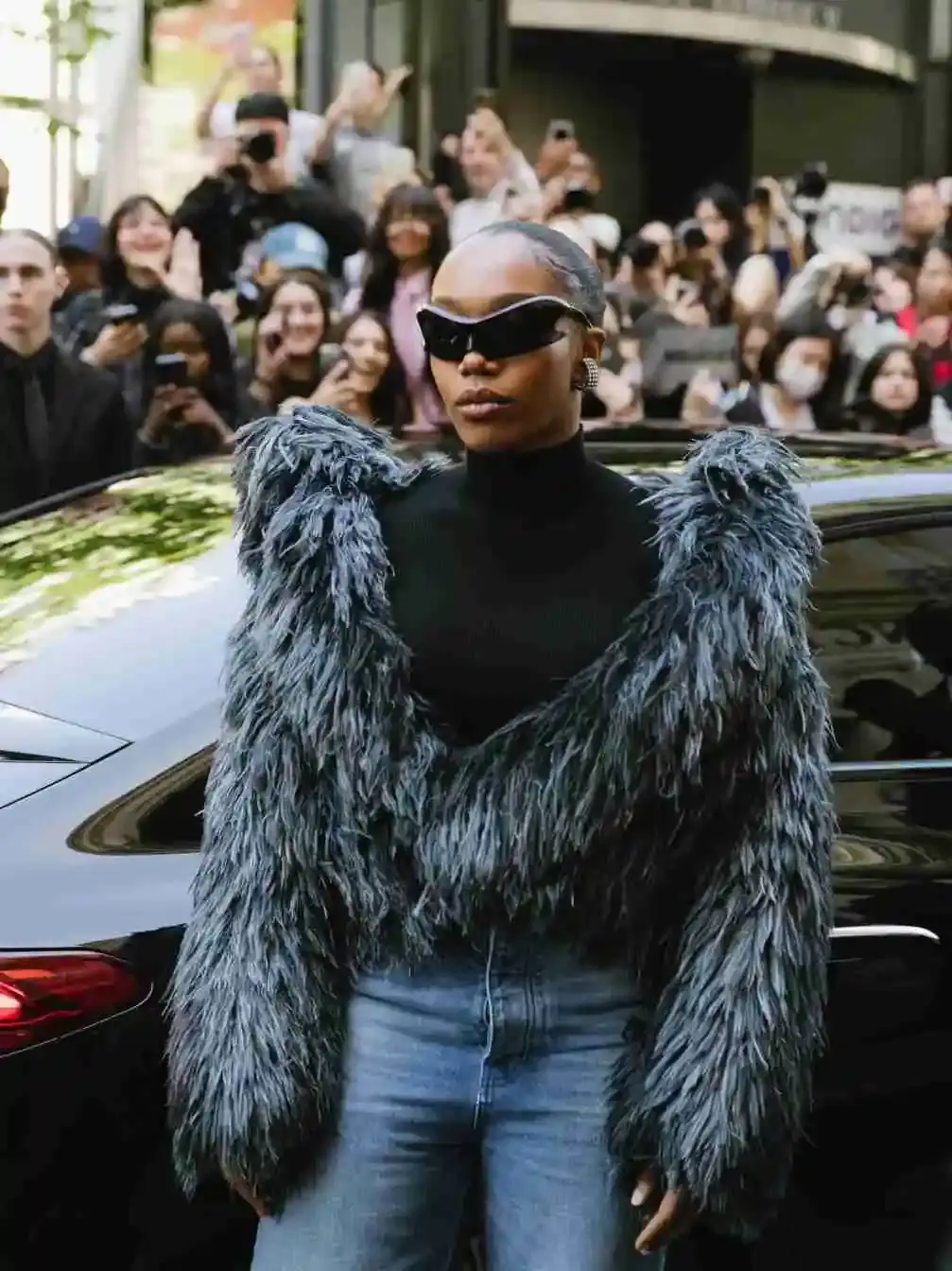 Naomi Ackie wearing a fluffy, grey/blue bolero over a black turtleneck and jeans, with futuristic sunglasses, surrounded by a crowd.