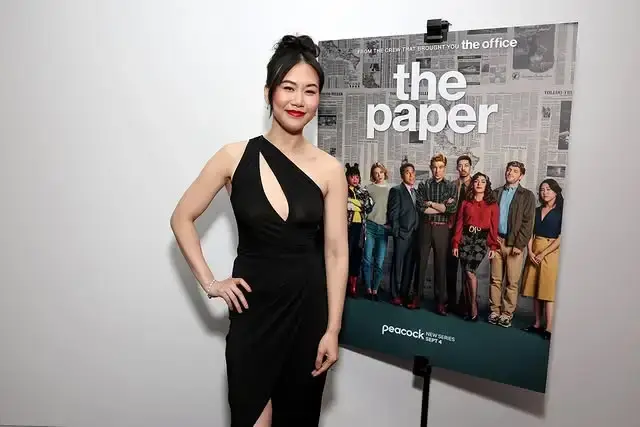 Actress Ramona Young wearing a black one-shoulder dress while posing next to a promotional poster for "The Paper."