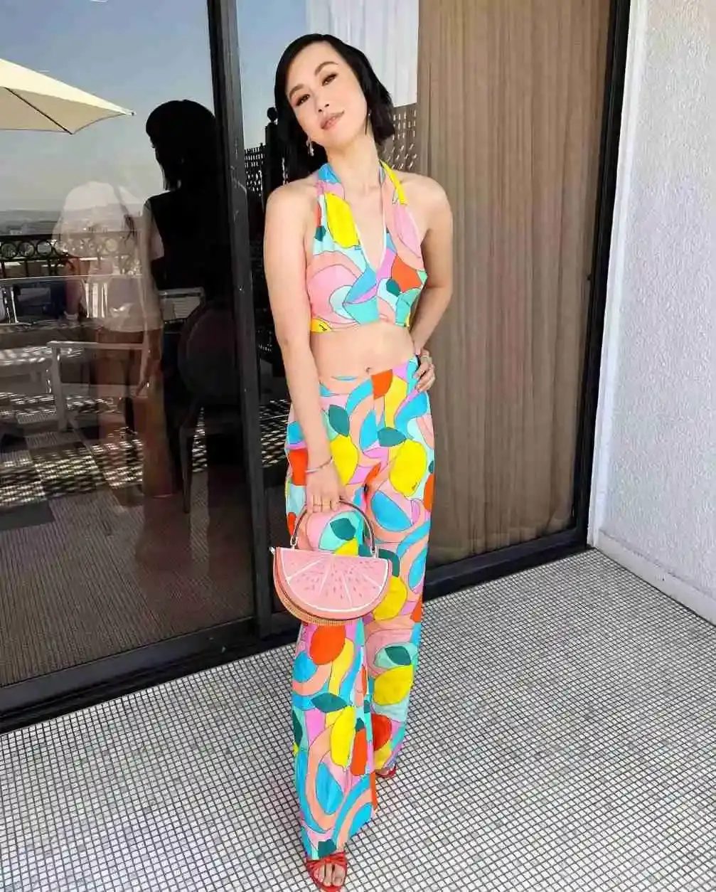 Full-body shot of actress Ramona Young wearing a colorful, abstract-print halter top and matching wide-leg pants, holding a watermelon-slice handbag.
