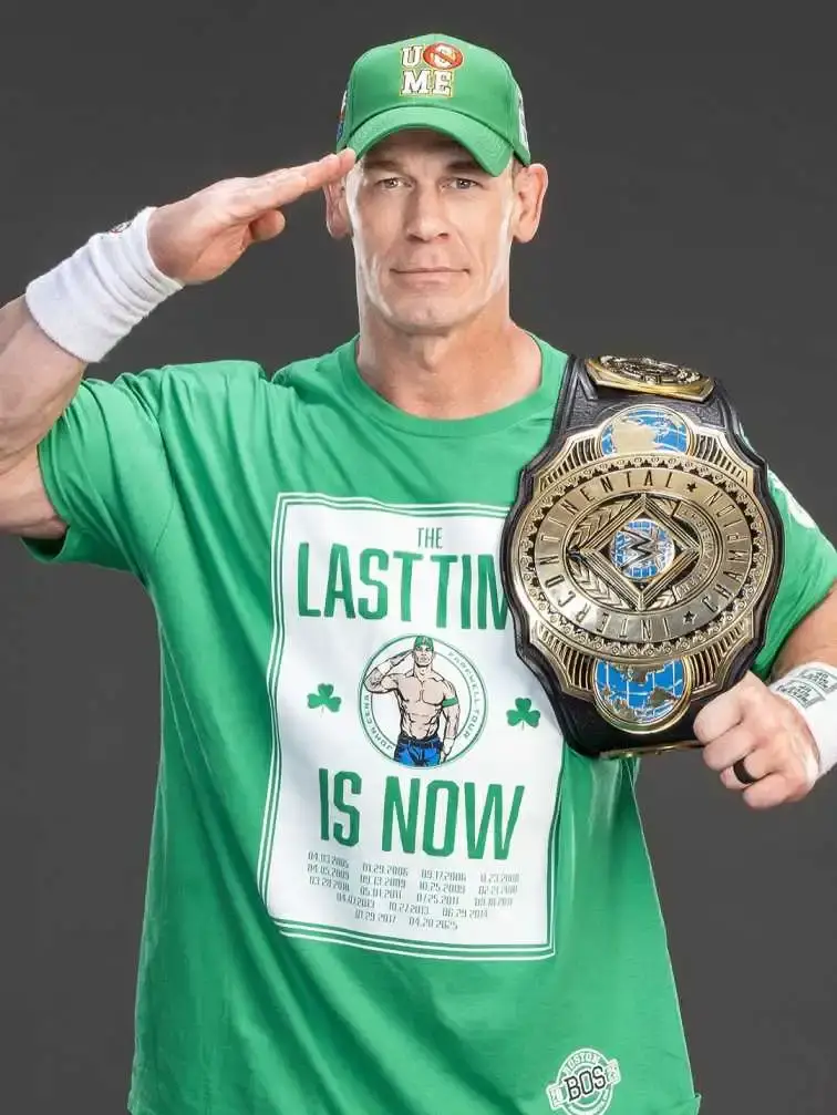 John Cena in a green "The Last Time Is Now" T-shirt and cap, holding the WWE United States Championship belt while saluting the camera.