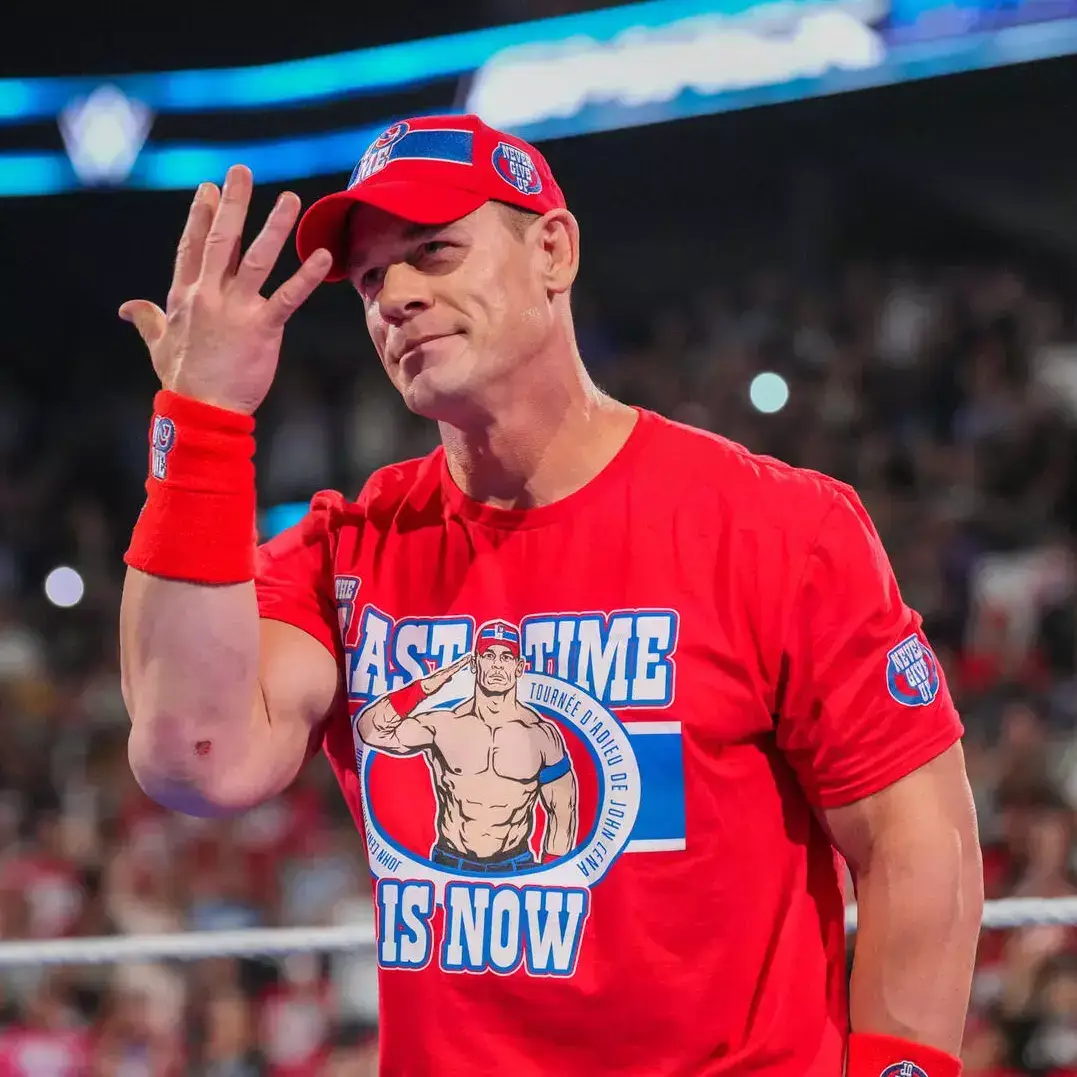 John Cena in the center of a wrestling ring, wearing a red "Last Time Is Now" T-shirt and cap, raising his hand to salute the crowd.