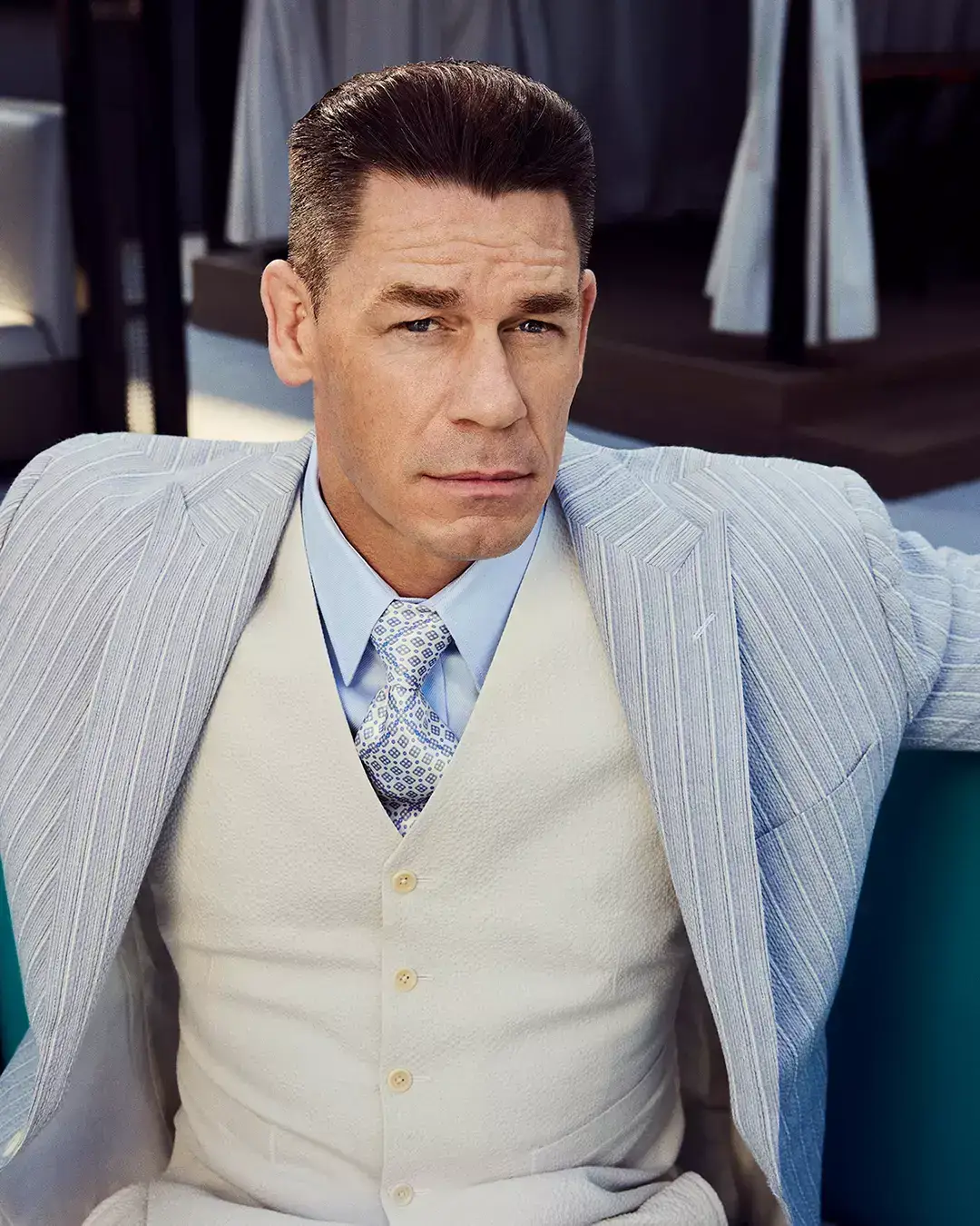 John Cena in a light blue pinstripe suit jacket over a white vest and light blue shirt with a patterned tie.