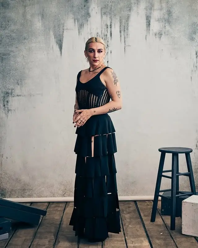 Mason Alexander Park posing indoors in a black tiered dress with a mesh cutout bodice, with visible arm tattoos and short blonde hair.