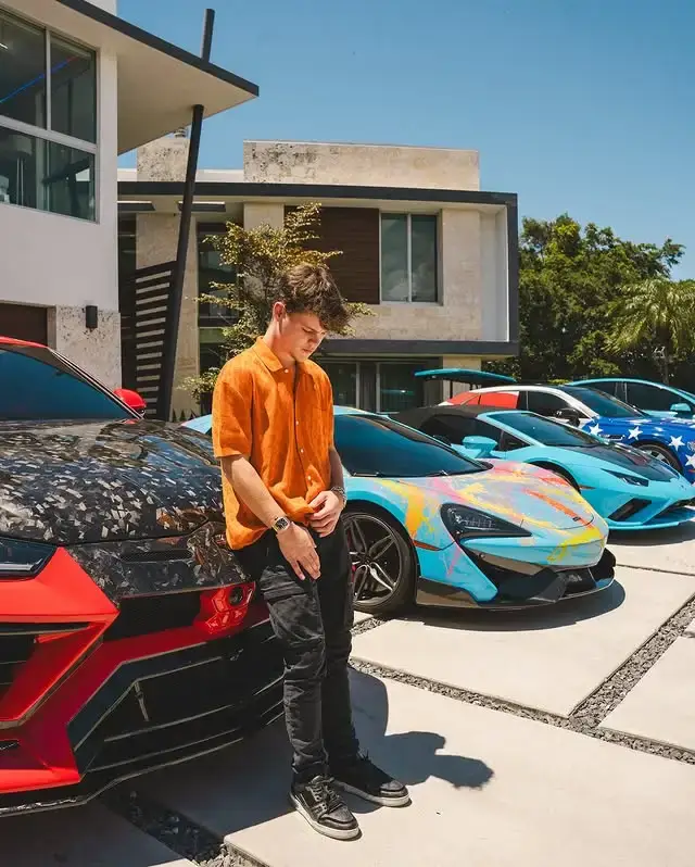 Jack Doherty leaning against a custom-wrapped red and black Lamborghini, with a row of other colorful supercars parked behind him outside a modern house.