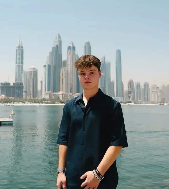 Jack Doherty standing by a body of water, wearing a short-sleeved navy blue button-up shirt and bracelets, with the skyscrapers of Dubai in the background.