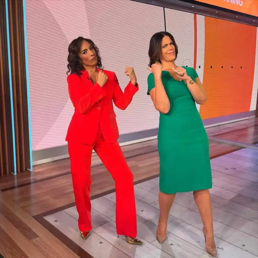 Michelle Miller in a red pantsuit and Dana Jacobson in a green dress stand on a TV set, both making playful fist-pumping gestures toward the camera.