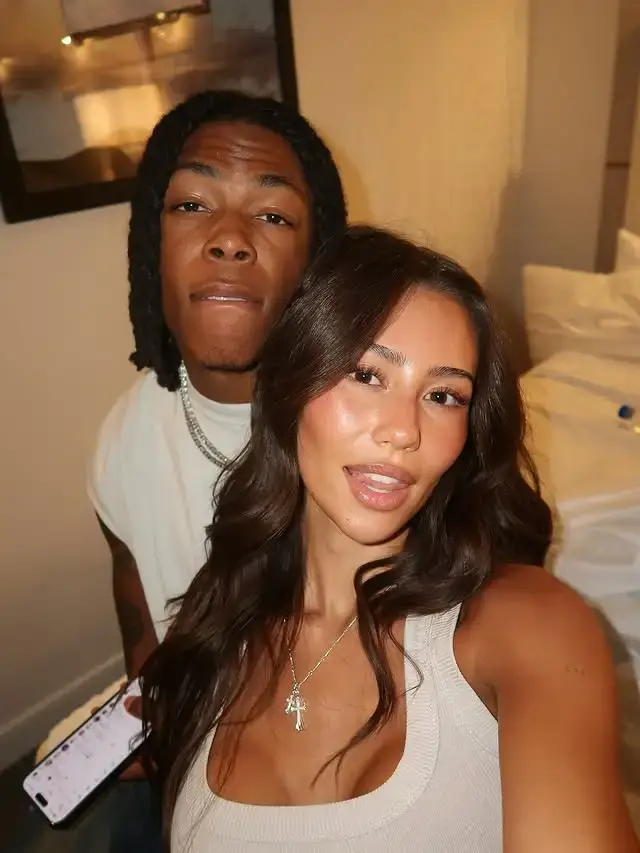 NFL player Jahmyr Gibbs, with long braids, stands behind his girlfriend Nicole Anderson, who wears a light tank top and cross necklace, as they take a selfie together.