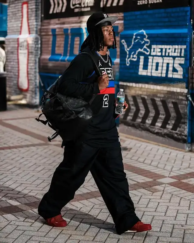 NFL player Jahmyr Gibbs walks on a brick sidewalk wearing all black, including a baseball cap, a long-sleeve jersey, baggy pants, and red sneakers, while carrying a large duffel bag and a water bottle.