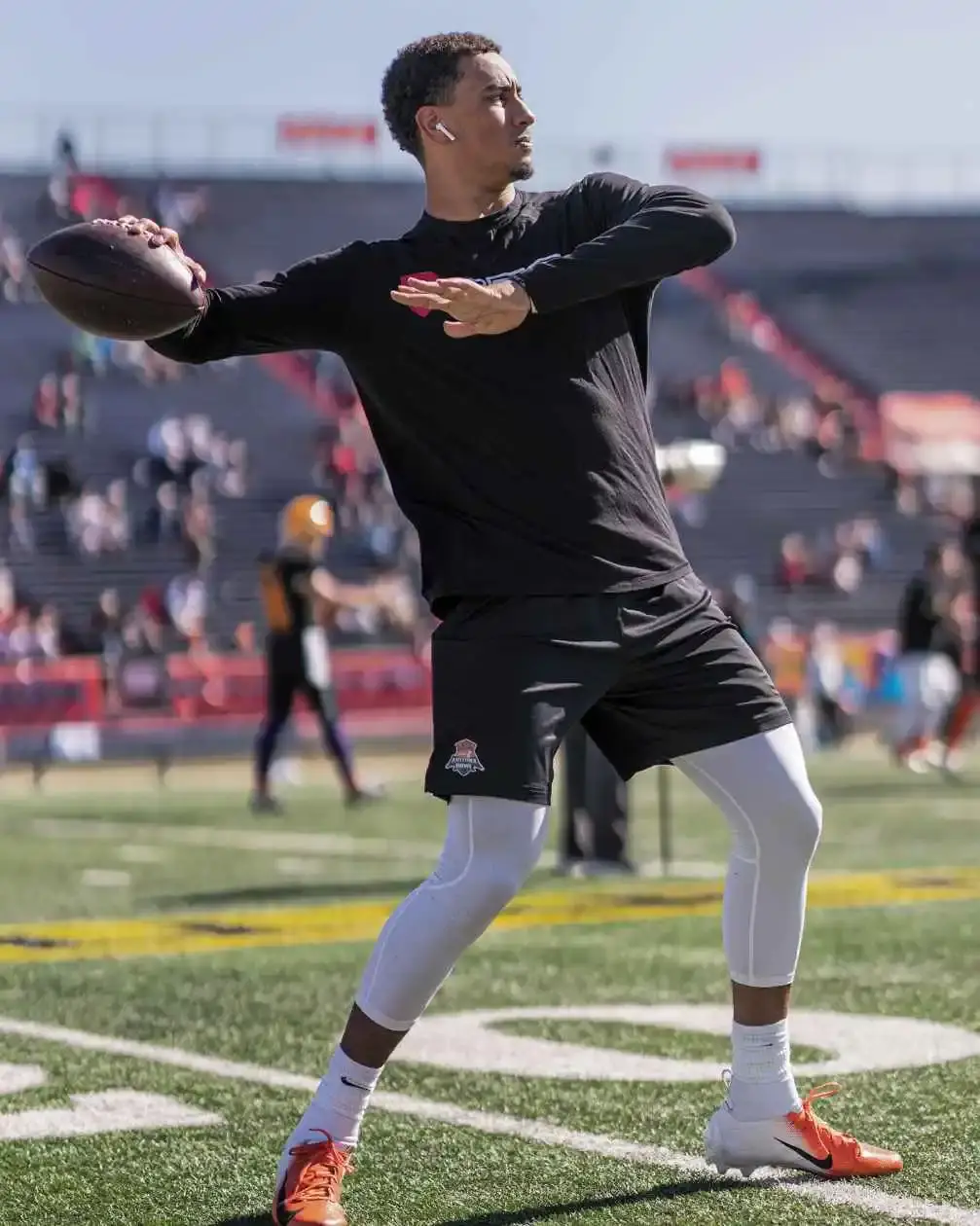 NFL quarterback Jordan Love throwing a football during an outdoor practice or warm-up, wearing a black long-sleeve shirt, black shorts, and white compression leggings.