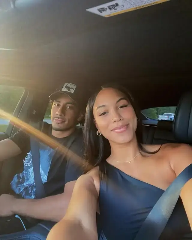 Jordan Love, wearing a blue T-shirt and a cap, and his wife, Ronika Stone, in a blue dress, smile while posing together at their car.