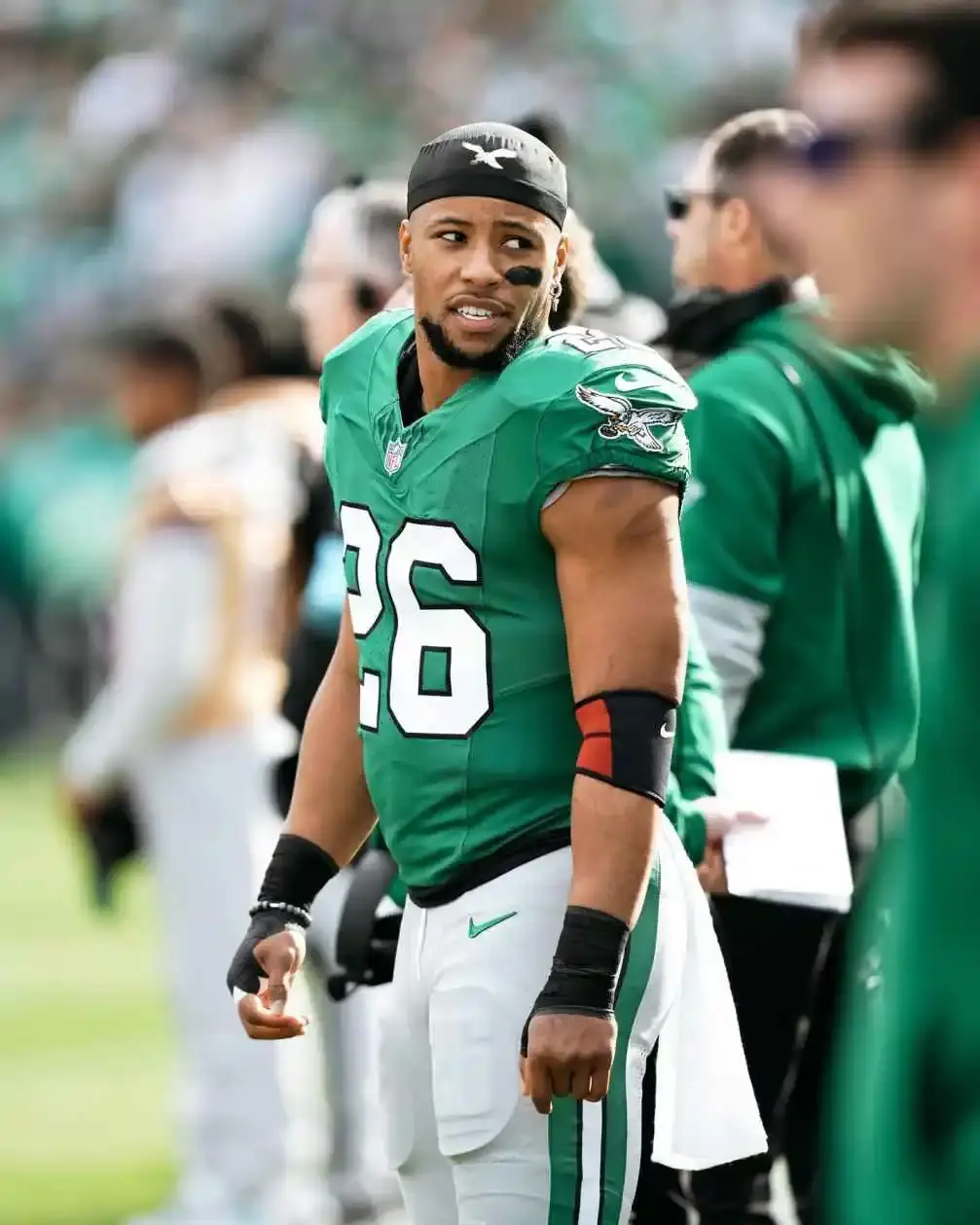 Saquon Barkley, wearing a green New York Jets jersey with the number 26, a black bandanna, and black under-eye paint, looks over his shoulder on the sidelines.