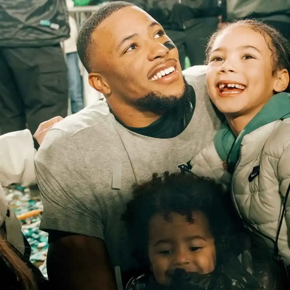 Saquon Barkley, smiling and looking up, kneels next to his happy-looking daughter, Jada Clare, and son, Saquon Jr., on a confetti-strewn field.