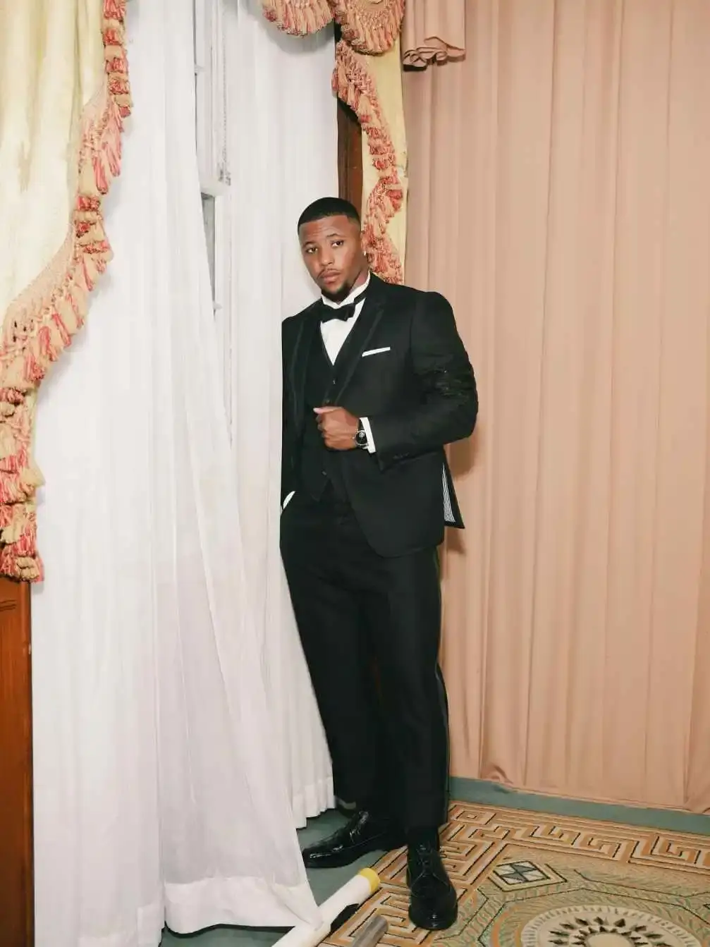 Saquon Barkley in a black tuxedo with a bow tie and vest, posing next to a window with white curtains.
