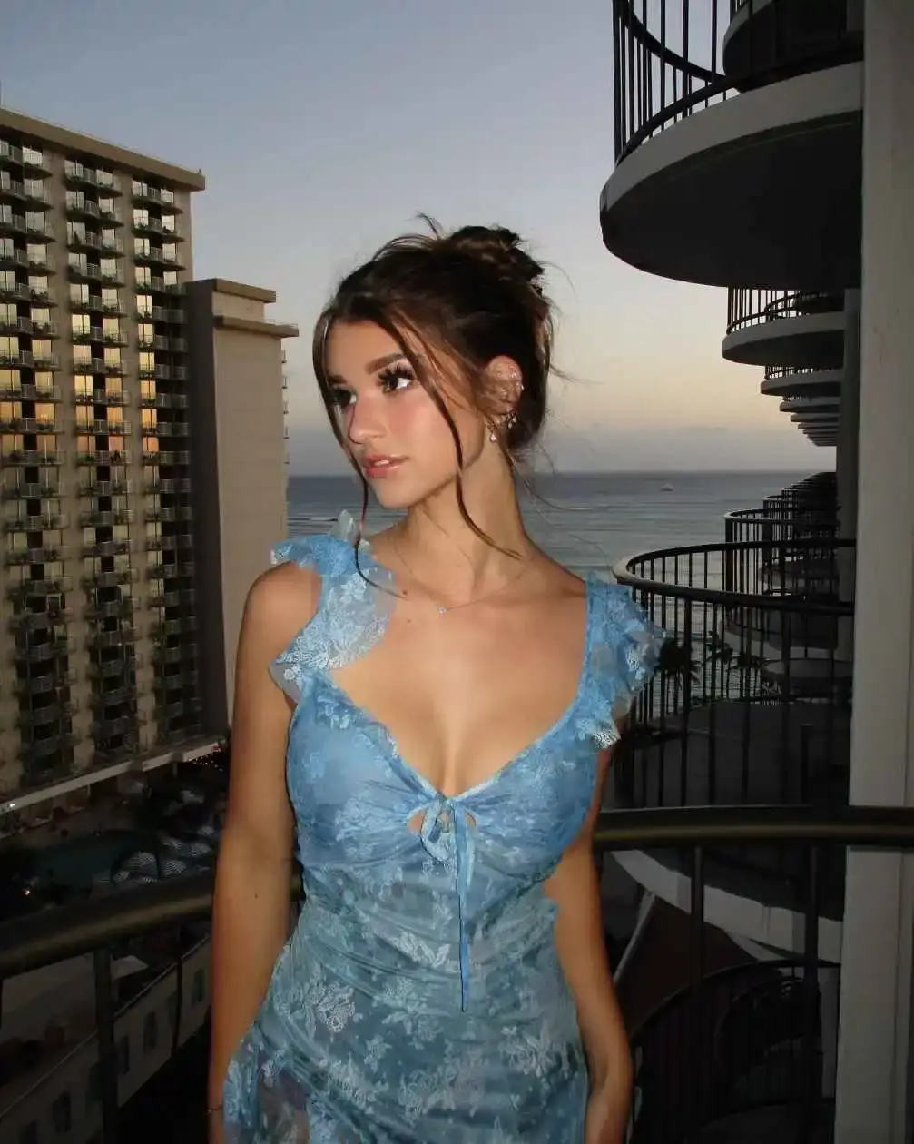 Brooke Monk standing on a balcony in a blue dress with a high-rise building and the ocean in the background at sunset.