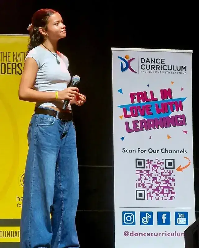 Catherine Laga’aia on stage holding a microphone, standing next to a banner for Dance Curriculum.