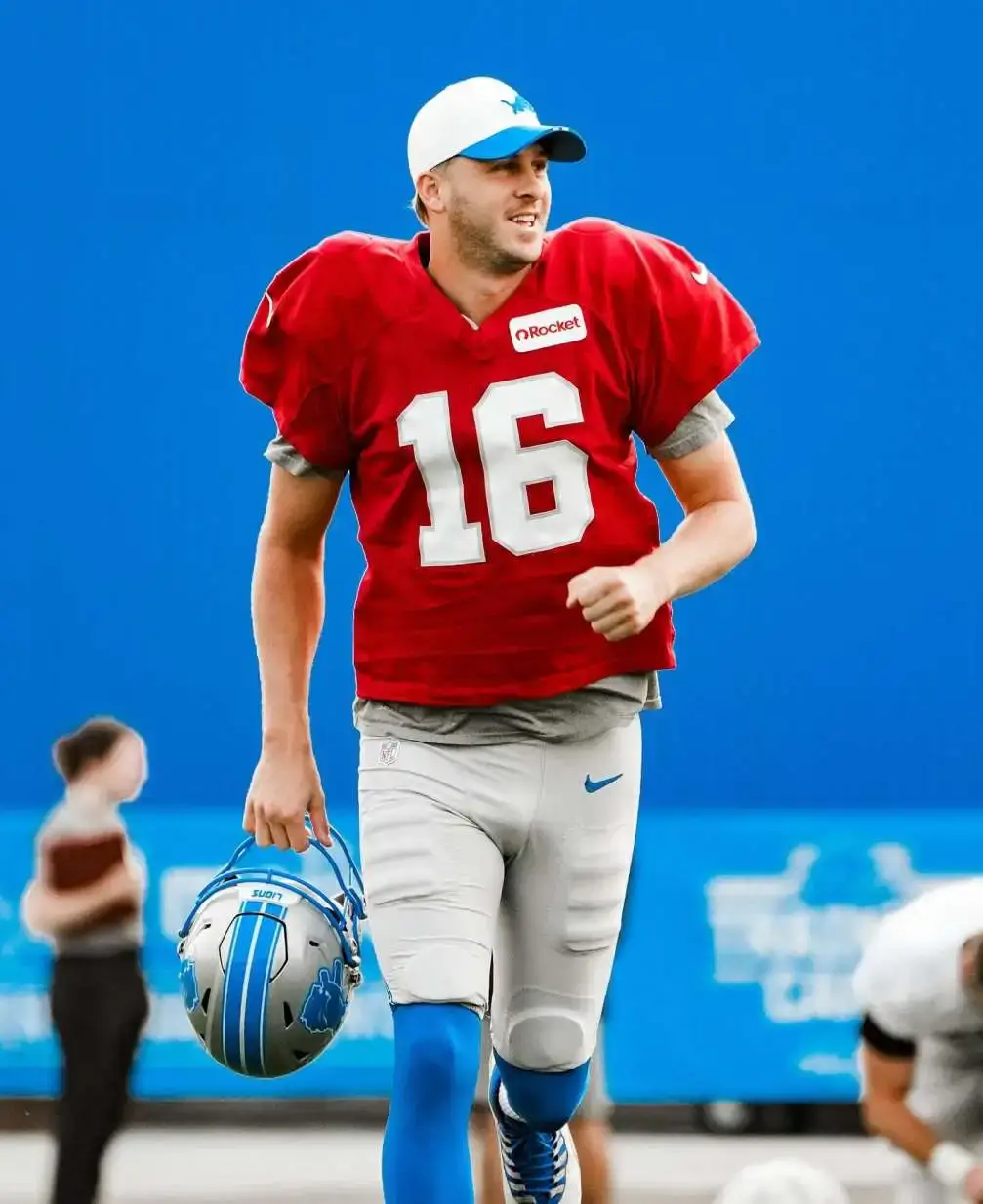 NFL quarterback Jared Goff wearing a Detroit Lions red practice jersey with the number 16, a white hat, and holding his silver and blue Lions helmet against a bright blue background.