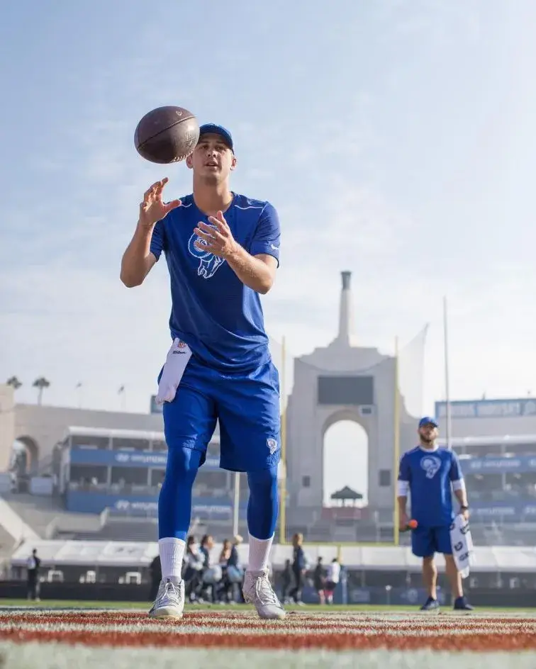 Quarterback Jared Goff in a blue Los Angeles Rams practice outfit catching a football high above a field with a stadium structure and archway visible in the background.