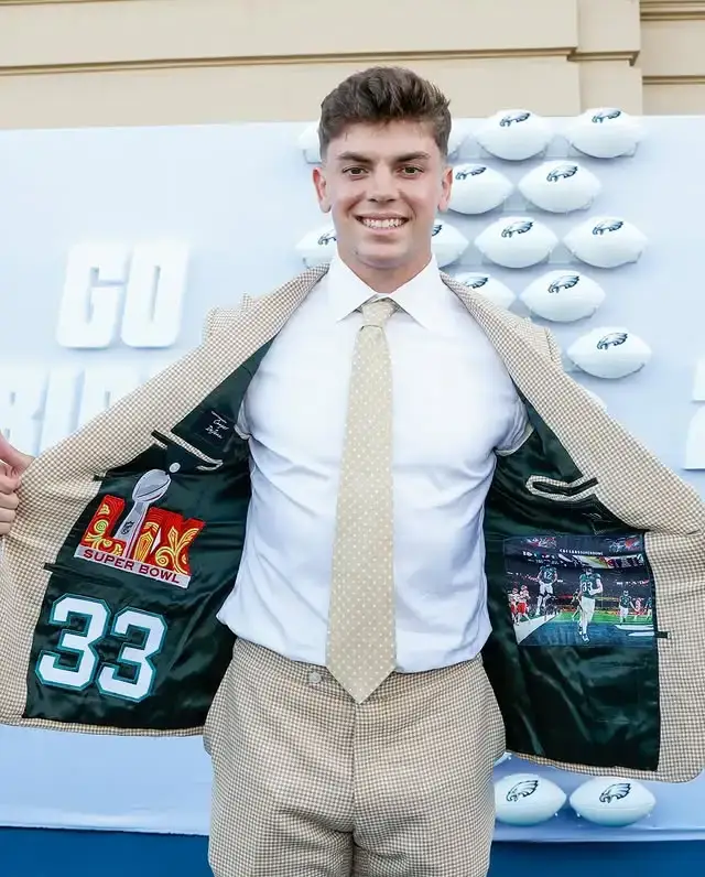 Cooper DeJean in a tan plaid suit, opening his jacket to reveal a black lining decorated with a Super Bowl trophy, the number 33, and football action photos.
