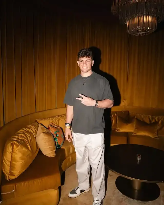 Cooper DeJean standing in a dimly lit, luxurious room with mustard yellow velvet seating and curtains, making a peace sign gesture.