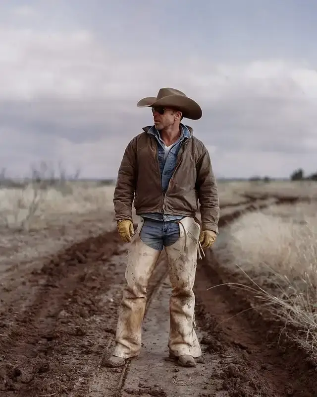 Screenwriter and director Taylor Sheridan standing outdoors in a muddy dirt track, wearing a brown jacket, light-wash jeans, chaps, sunglasses, and a wide-brim cowboy hat.