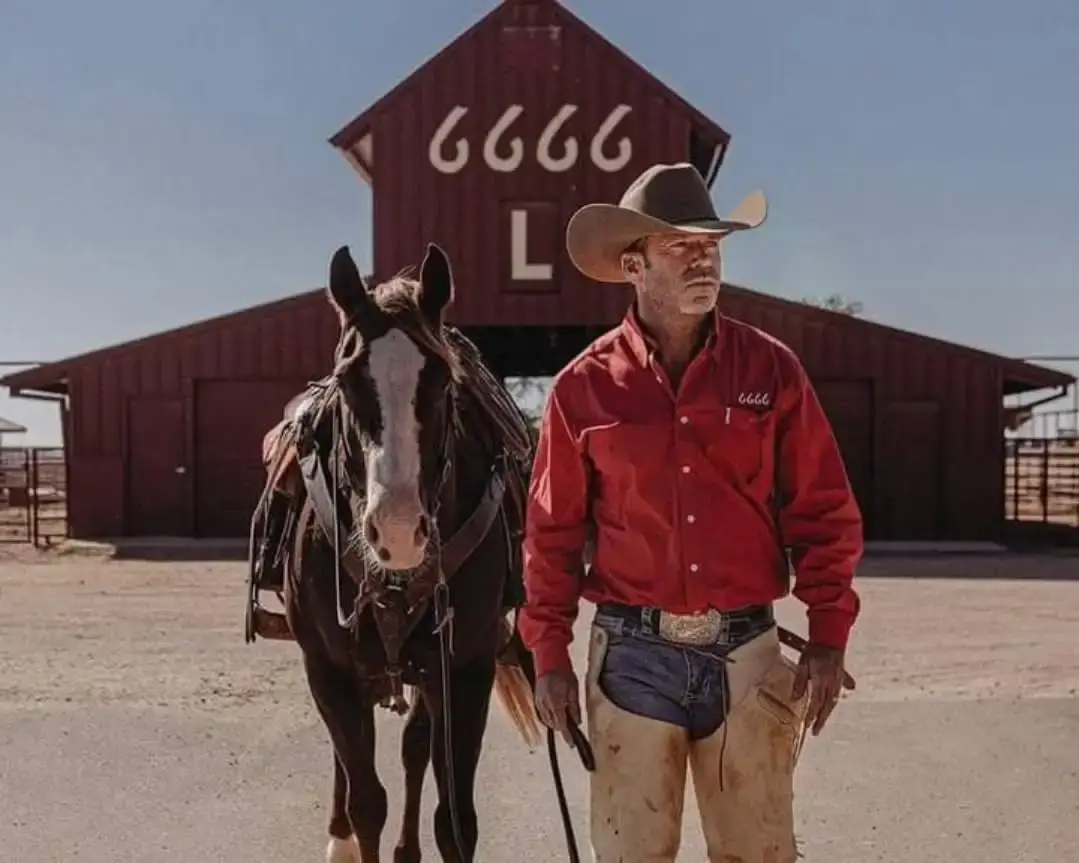 Taylor Sheridan standing next to a dark brown horse, wearing a red long-sleeve Western shirt and chaps, in front of a red barn building marked with "6666 L" on a sunny day.